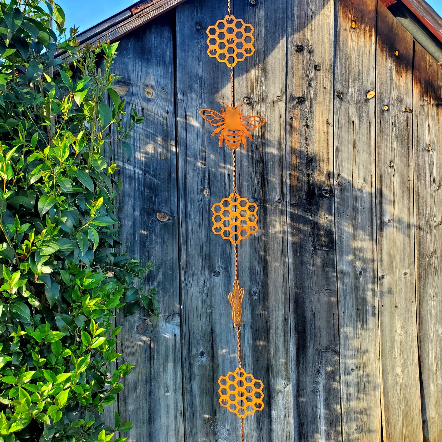 Bee and honeycomb wind spinner made of rusted steel, close-up view showing intricate laser-cut bee with wings and hexagonal honeycomb patterns against a garden shed. 