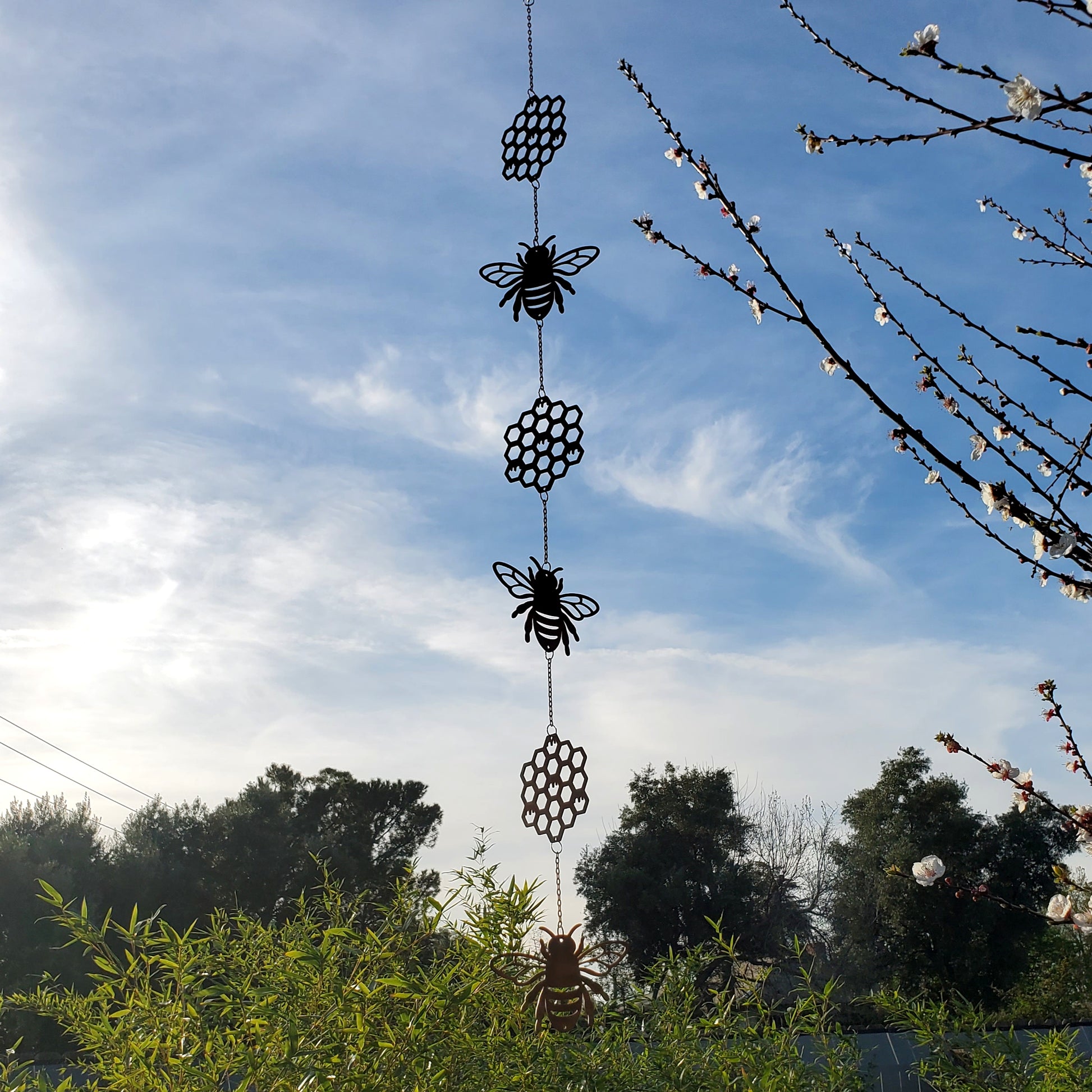 Decorative hanging wind spinner with bees and honeycomb patterns against a blue sky with trees and flowers.