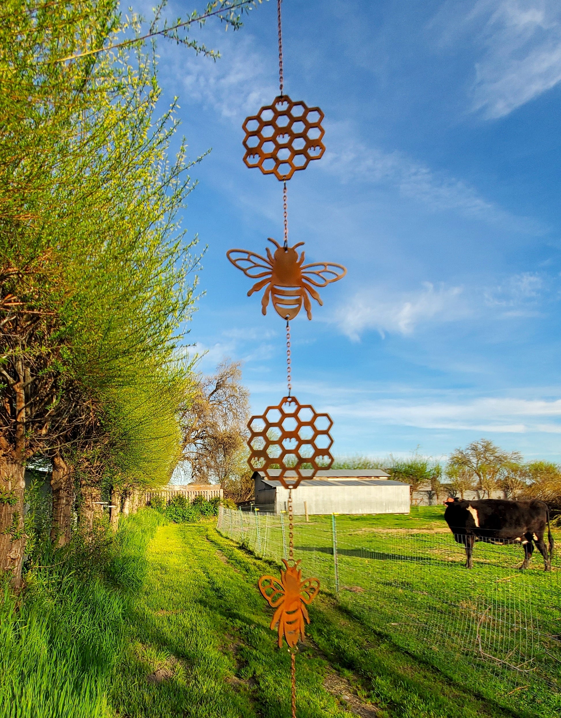 Decorative metal bee and honeycomb wind chime against a blue sky with trees and a cow in the background.