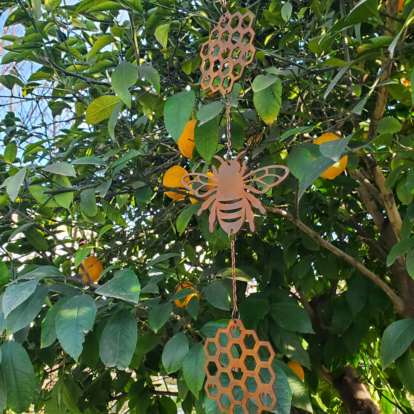 Decorative bee-shaped windchime  hanging from a tree with green leaves and lemon  fruits.